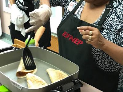 Two women wearing EFNEP aprons cooking folded tortillas on electric griddle