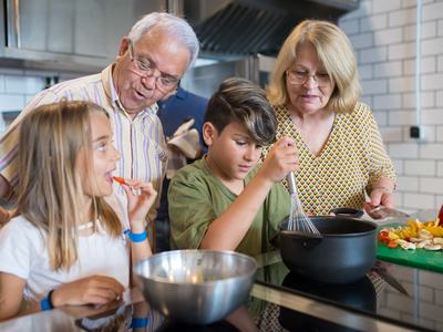 Grandparents with two children cooking; boy whisking a pot, girl tasting a pepper