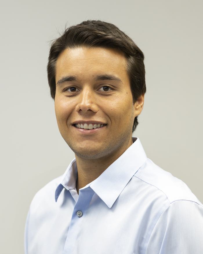 Young man in light blue dress shirt, headshot on plain gray background