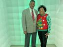 Man in gray suit with red striped tie and woman in red Christmas sweater in green-lit inflatable booth