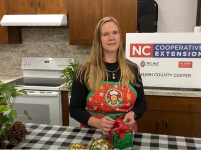 Woman in holiday apron wrapping a gift in a kitchen; sign reads "NC Cooperative Extension"