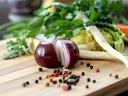 Halved red onion with peppercorns on cutting board, herbs and root vegetables behind