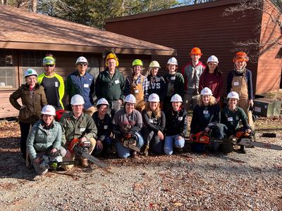 Group of people wearing hard hats and holding chainsaws in front of a wooden building