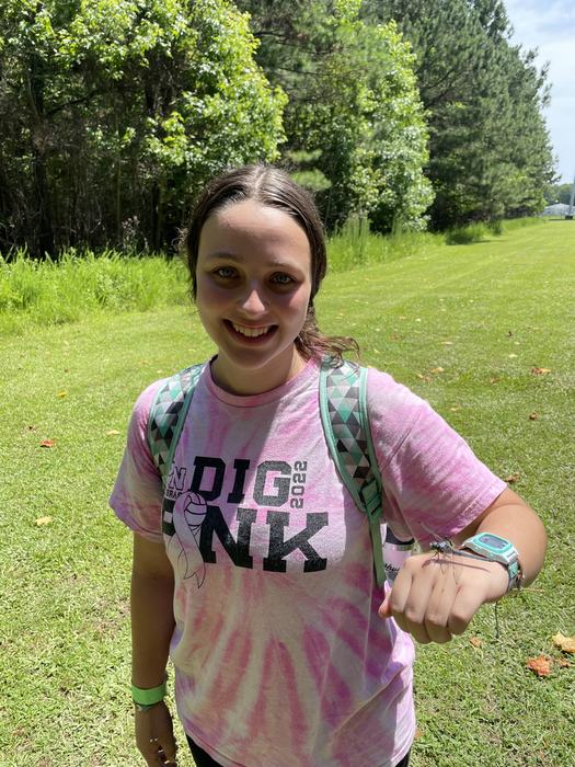 Young person holding a dragonfly on their hand, wearing tie-dye shirt reading "DIG PINK 2022"