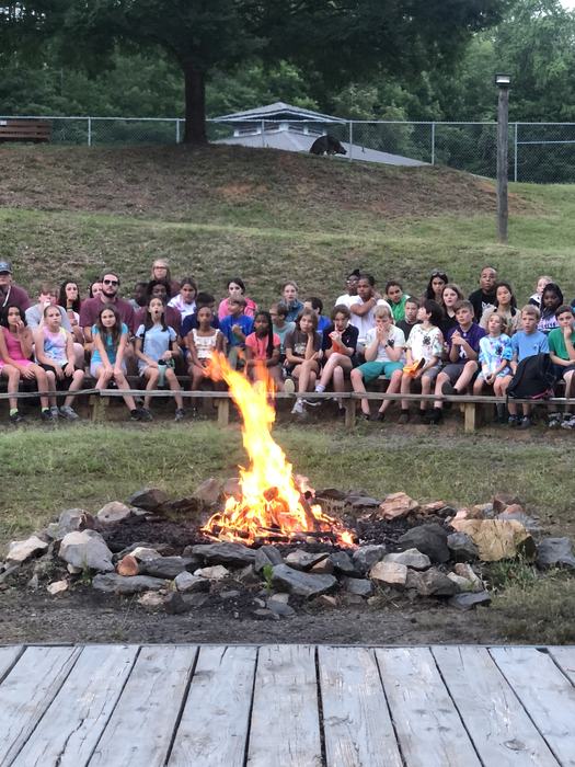 Campfire burning in a stone firepit with a group of children and adults seated on benches