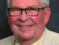 Older man with white hair and glasses wearing beige suit, yellow shirt, patterned tie, U.S. flag pin