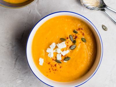 Bowl of orange soup garnished with feta, pumpkin seeds, and paprika, spoon nearby