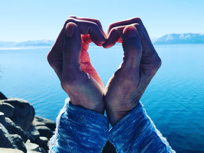 Two hands forming a heart shape framing a blue lake and distant mountains
