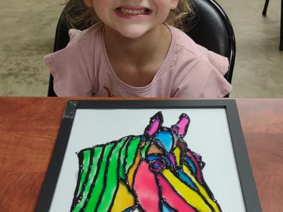 Young child sitting behind a framed, colorful painted horse head artwork on a table