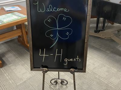Chalkboard on easel reading "Welcome" and "4‑H guests" with a drawn four-leaf clover