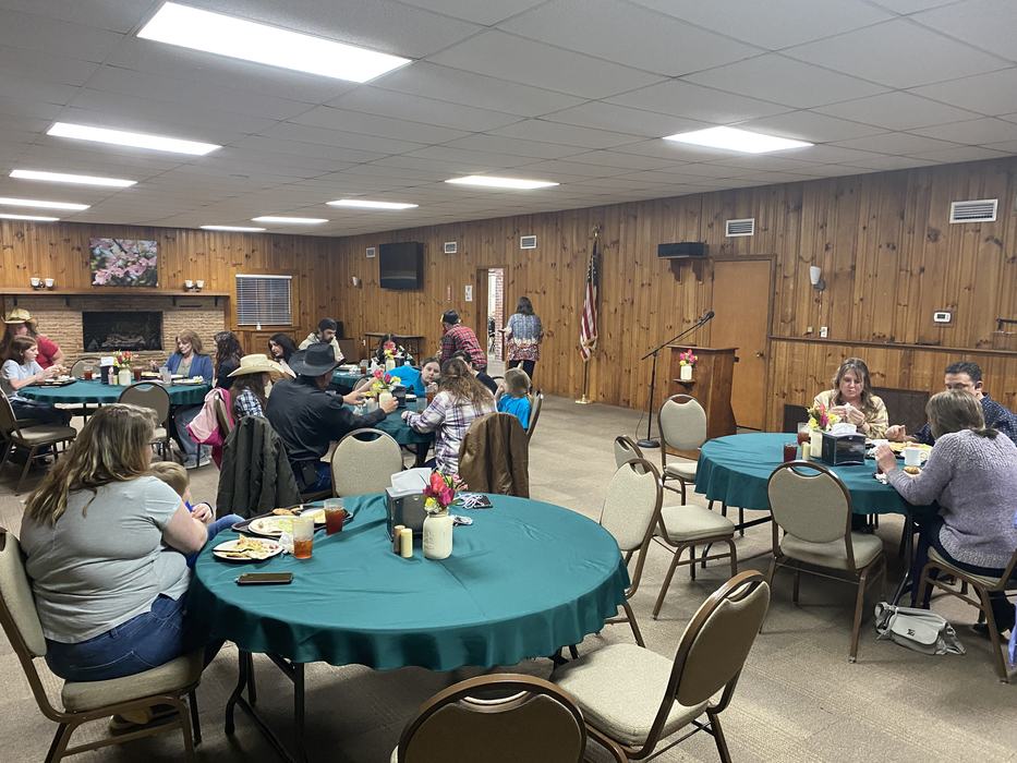 Community members seated at round tables eating in a wood-paneled hall with podium and flag