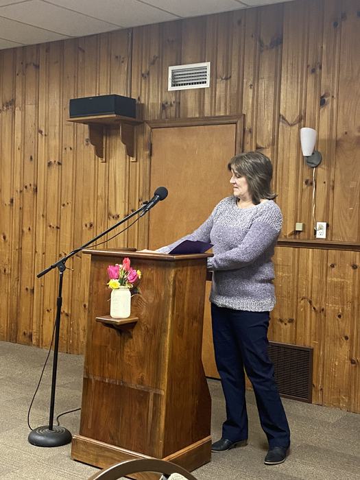 Person standing at wooden podium with microphone and jar of flowers