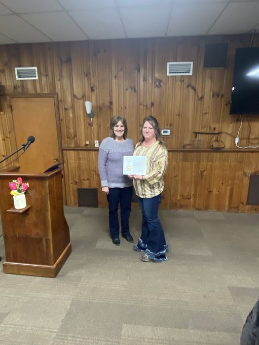 Two women standing in front of wood-paneled wall, one holding a framed certificate, podium left