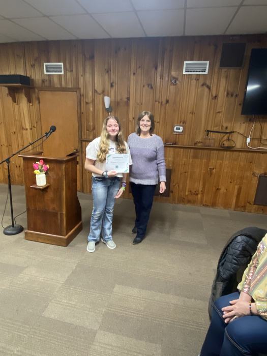 Teen holding certificate standing next to adult by podium in wood-paneled room