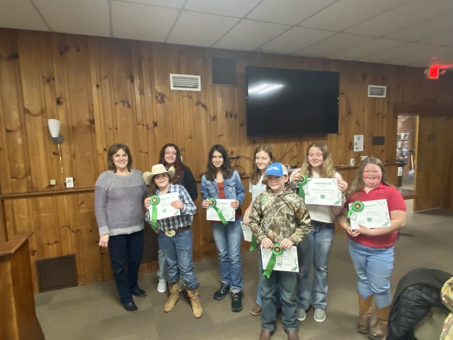 Group of children and an adult holding 4-H certificates and green ribbons in wood-paneled room