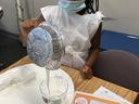 Girl wearing mask pours white liquid from foil pan into measuring cup over plate