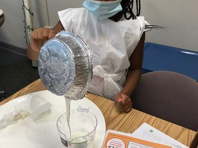 Girl wearing mask pours white liquid from foil pan into measuring cup over plate