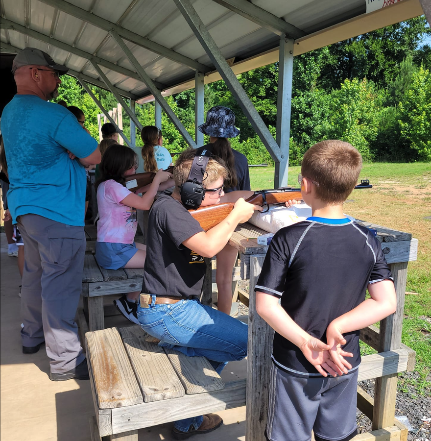 Child with ear protection aiming a rifle at a shooting-range bench outdoors
