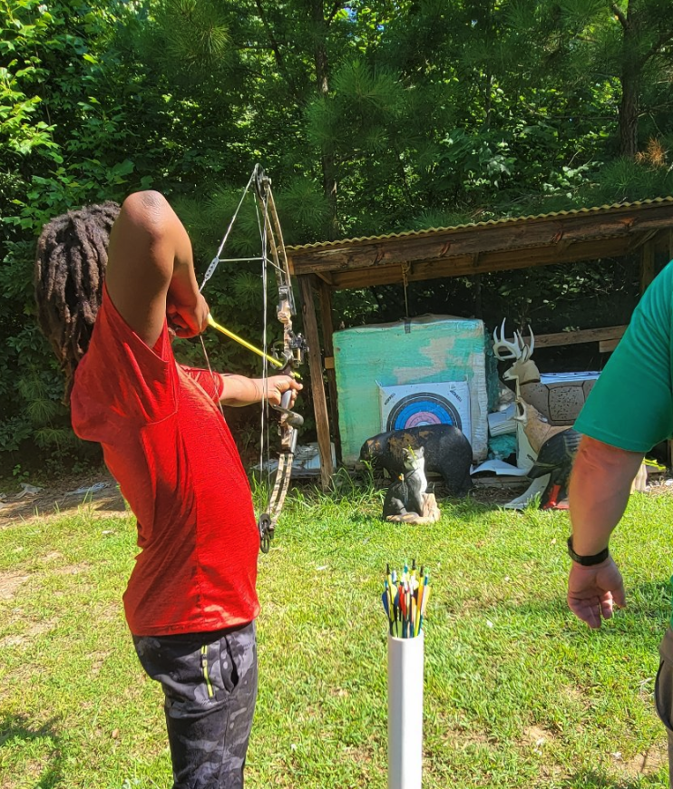 Person in red shirt drawing a bow toward a paper target at an outdoor archery range
