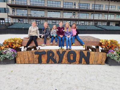Six children sitting on a wooden beam above a sign reading "TRYON" at an outdoor arena