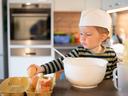 Young child in a chef hat reaching from an egg carton toward a large mixing bowl