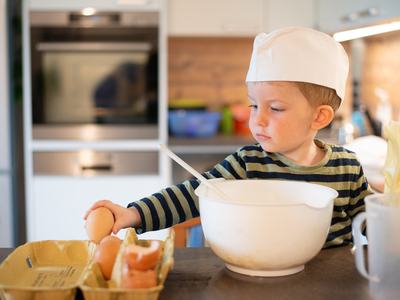 Young child in a chef hat reaching from an egg carton toward a large mixing bowl