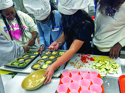 Group of youth in chef hats filling muffin tins with vegetable mixtures and sliced zucchini