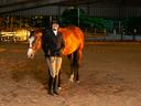 Woman in riding attire holding reins of a chestnut horse inside covered arena.