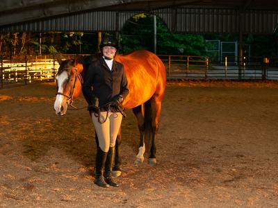Woman in riding attire holding reins of a chestnut horse inside covered arena.
