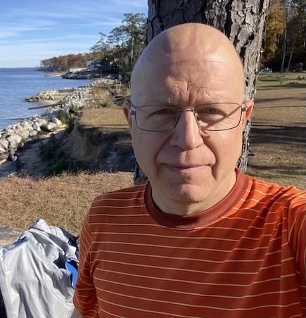 Bald man with glasses in orange striped shirt selfie by lake and rocky shore