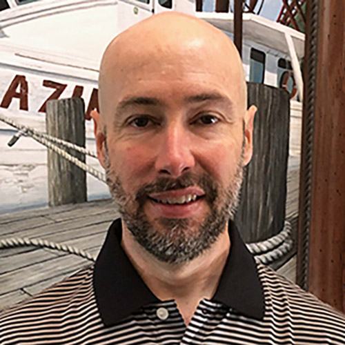 Bald man with short beard in striped polo standing before dock and boat mural showing "AZU"