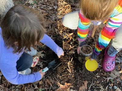 Two children crouched on leaf-covered ground digging soil with a trowel and a jar