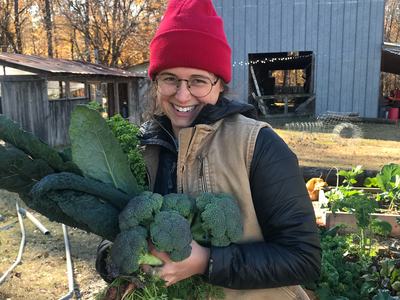Person in red beanie holding broccoli, carrots, and kale in a garden