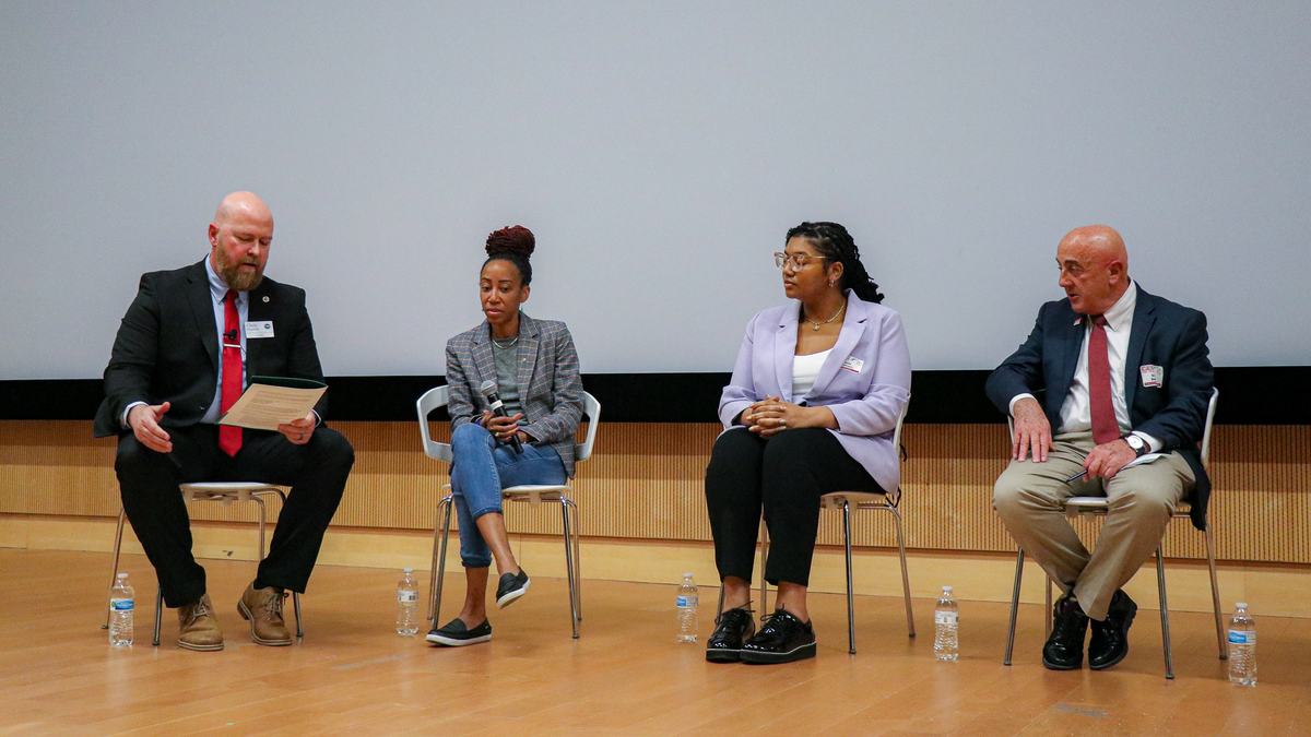 Four seated panelists on stage during a discussion; one holds papers, one a mic.