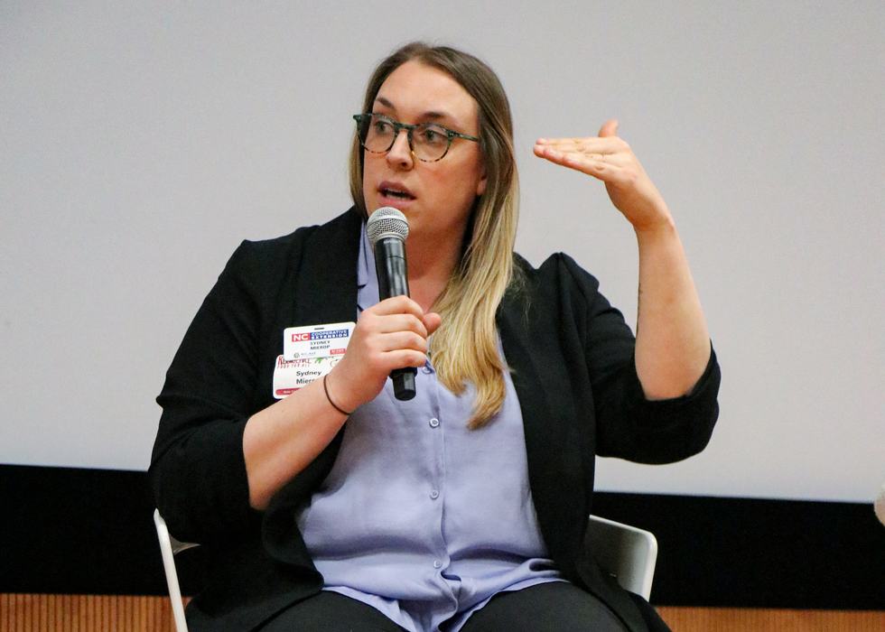Person holding a microphone and gesturing with right hand, wearing a name tag with printed text.