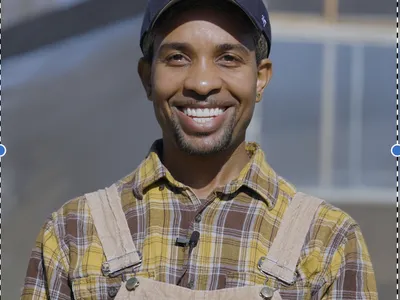 Person wearing cap, plaid shirt, and overalls with clip-on microphone outdoors