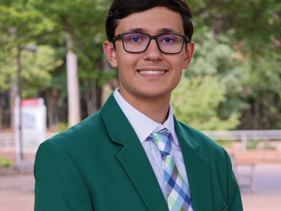 Young man wearing a green blazer, plaid tie and glasses, standing outdoors