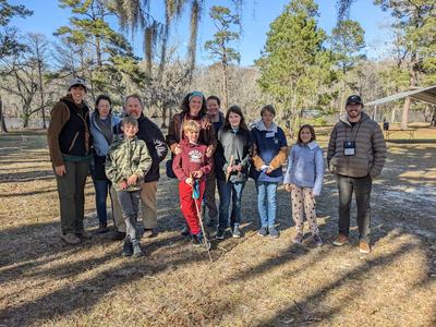 Group of adults and children standing outdoors in a wooded park clearing