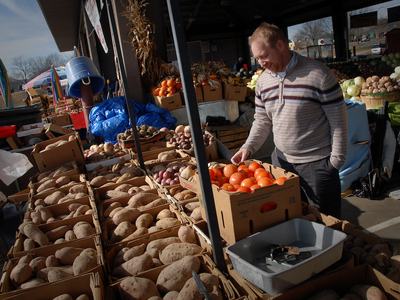 Man at outdoor market picking a tomato from a box beside boxes of potatoes