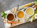Three bowls of broths on a cutting board surrounded by vegetables and herbs