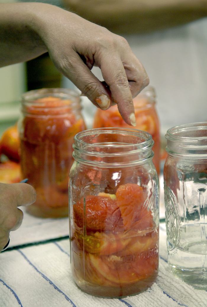 A hand points down into a jar of tomatoes being prepared for canning.