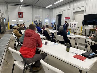 People seated at tables listening to a presenter delivering a workshop inside a warehouse facility