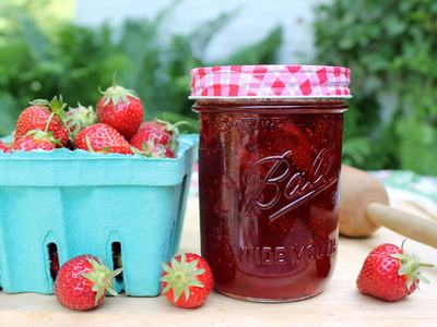 Mason jar of strawberry jam labeled "Ball WIDE MOUTH" beside a carton of fresh strawberries
