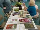 Two women at a table displaying gardening books and potted plants in a gymnasium