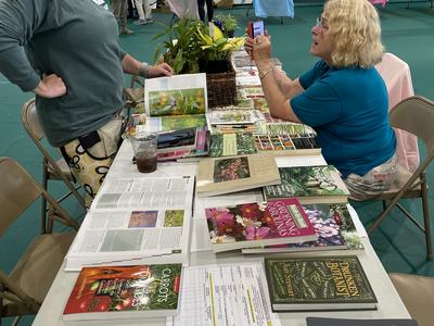 Two women at a table displaying gardening books and potted plants in a gymnasium