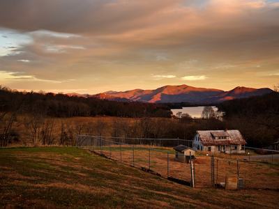 Rusted-roof farmhouse by fenced yard with lake and mountains at sunset