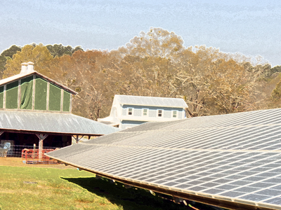 Ground-mounted solar panel array in front of farm buildings and wooded hillside