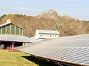 Ground-mounted solar panel array in front of farm buildings and wooded hillside