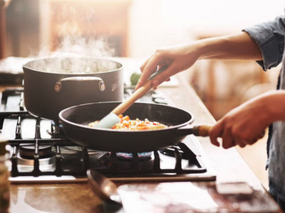 Hands stirring vegetables in a frying pan on stovetop with a steaming pot nearby