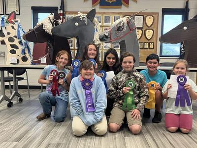 Eight children kneeling holding 4-H ribbons in front of large horse-head sculptures